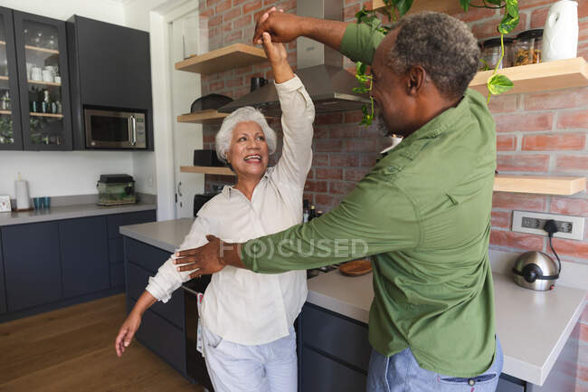 Щаслива старша афроамериканська пара у відставці, яка тримається за руки, танцює разом і посміхається на кухні, і ізолюється вдома під час коронавірусної пандемії. — стокове фото