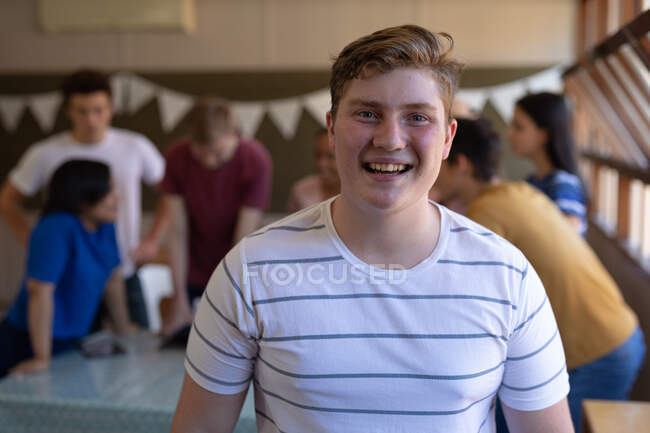Porträt eines kaukasischen Teenagers mit kurzen Haaren und grauen Augen, der in einem Klassenzimmer steht und in die Kamera lächelt, während sich Mitschüler im Hintergrund unterhalten — Stockfoto
