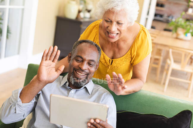 Счастливая пожилая афроамериканская пара дома, в своей гостиной, мужчина сидит на диване, держа планшетный компьютер и машет рукой, женщина стоит позади, глядя на планшетный компьютер вместе и улыбаясь — стоковое фото