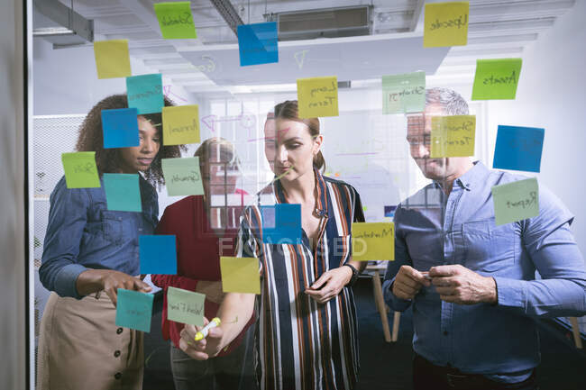 Un gruppo eterogeneo di colleghi d'affari che lavorano in un ufficio moderno, facendo brainstorming e scrivendo su una parete trasparente con appunti, visti attraverso il vetro — Foto stock
