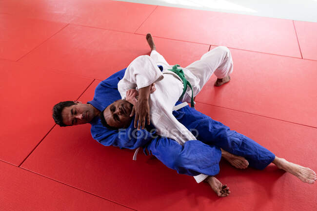Vista frontal de alto ângulo de uma raça mista judoca masculina de judo treinador e adolescente mista vestindo judoca azul e branca, praticando judô durante um treinamento em um ginásio. — Fotografia de Stock