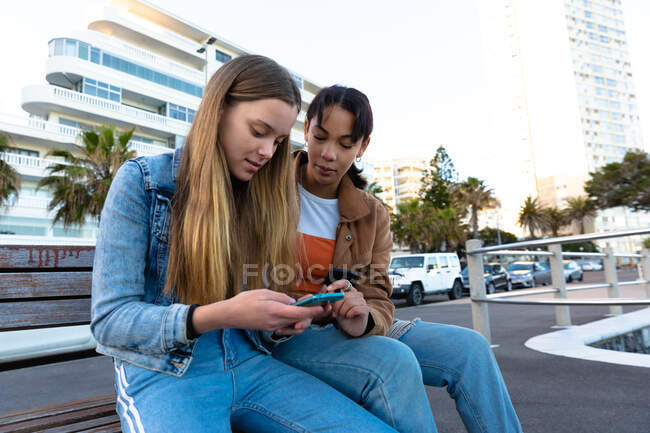 Передній вид на кавказьку і змішану расу дівчат, які проводять час разом в сонячний день, сидячи на лавці, дівчинка тримає смартфон, показуючи його своєму другу. — стокове фото
