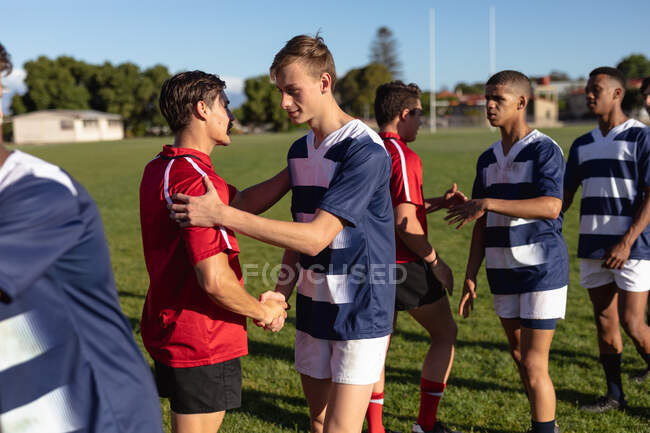 Seitenansicht von zwei multiethnischen Männerteams mit Rugby-Spielern, die ihre Mannschaftsstreifen tragen, sich auf dem Spielfeld grüßen, Hände schütteln und vor Spielbeginn lächeln — Stockfoto