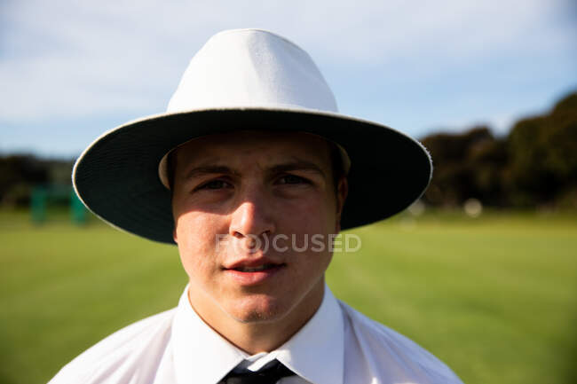 Портрет уверенного кавказского судьи в белой рубашке, черном галстуке и широкой шляпе с полями, стоящего на поле для крикета в солнечный день, смотрящего в камеру. — стоковое фото