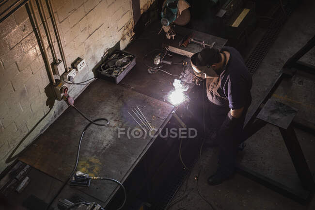 Caucasian male factory worker wearing dark apron and welding mask, standing at a workbench, welding. — Stock Photo