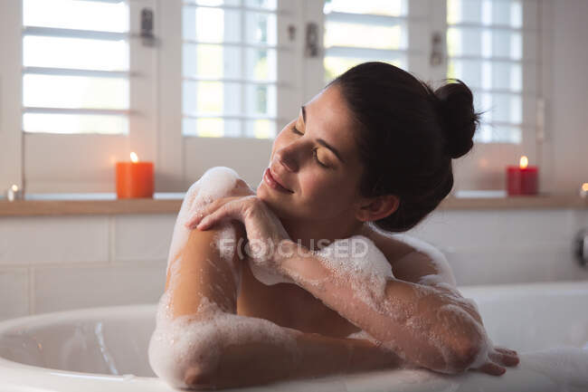 Женщина смешанной расы проводит время дома, лежа в ванне, расслабляясь с закрытыми глазами в ванной комнате. Самоизоляция и социальное дистанцирование в карантинной изоляции во время эпидемии коронавируса ковида 19. — стоковое фото