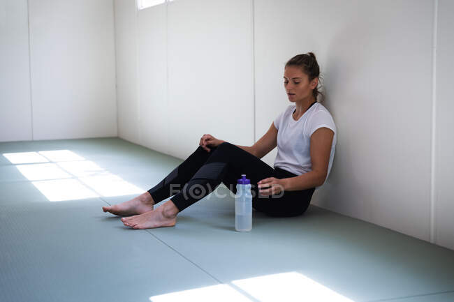Vista lateral de uma jogadora de judô caucasiana sentada no ginásio, fazendo uma pausa no treinamento, pegando uma garrafa de plástico de um tapete. — Fotografia de Stock