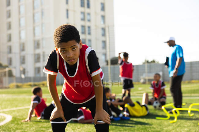 Vorderansicht eines Mischlingsfußballers, der mit den Händen auf den Knien auf einem Spielfeld steht und während des Trainings in die Kamera schaut, während seine Teamkollegen im Hintergrund ihrem Trainer zuhören. — Stockfoto
