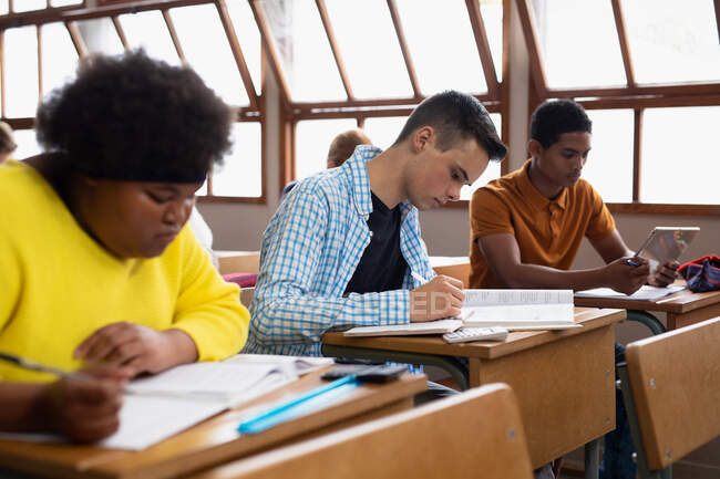 Visão lateral de um adolescente caucasiano em uma sala de aula da escola sentado na mesa, concentrando-se e escrevendo, com adolescentes mestiços masculinos e afro-americanos colegas de classe sentados em mesas de trabalho em cada lado dele — Fotografia de Stock