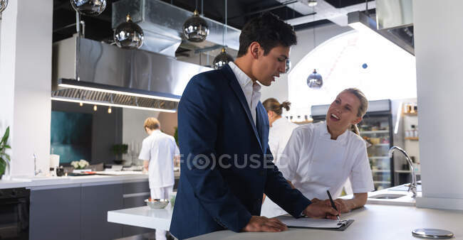Hombre de raza mixta vistiendo un traje, escribiendo en un archivo de papeles, hablando con una chef blanca, sonriendo, con otros cocineros cocinando en el fondo. - foto de stock