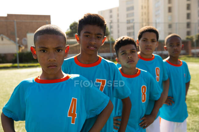 Frontansicht einer multiethnischen Gruppe von Jungen-Fußballern, die ihren blauen Mannschaftsstreifen tragen, an einem sonnigen Tag in Formation auf einem Spielfeld stehend, die Hände auf den Hüften, direkt in die Kamera blickend — Stockfoto