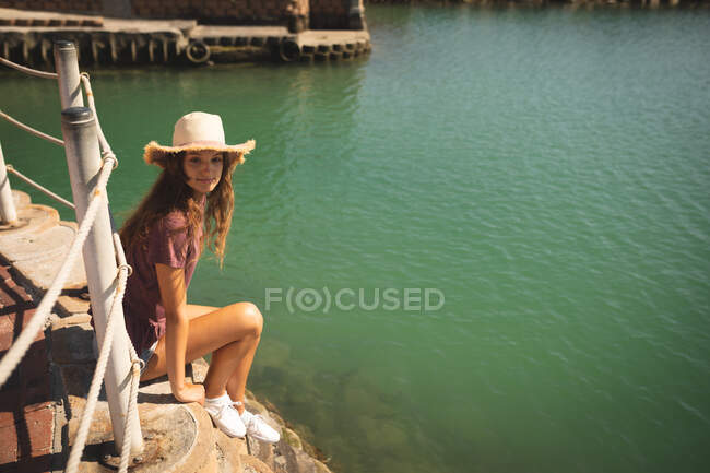 Una adolescente caucásica, usando un sombrero de paja, disfrutando de su tiempo en un paseo marítimo, en un día soleado, sentada detrás de una barrera, mirando hacia otro lado - foto de stock