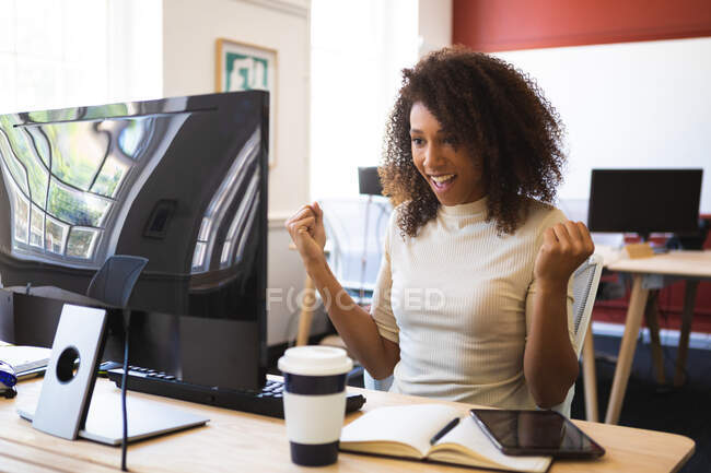 Femme d'affaires mixte aux cheveux bouclés, travaillant dans un bureau moderne, assise à une table et souriante, utilisant un ordinateur de bureau — Photo de stock
