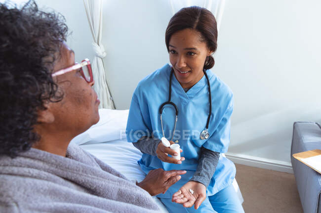 Femme âgée métisse passant du temps à la maison, étant visitée par une infirmière métisse, l'infirmière lui donnant des médicaments — Photo de stock