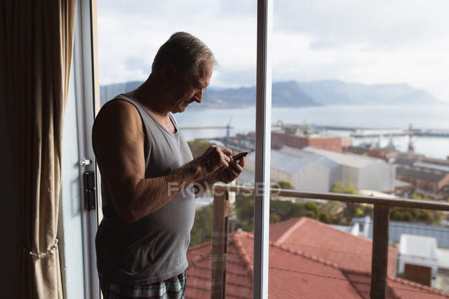 Vista lateral de un hombre caucásico mayor que se relaja en casa con un chaleco, parado junto a la ventana usando un teléfono móvil - foto de stock