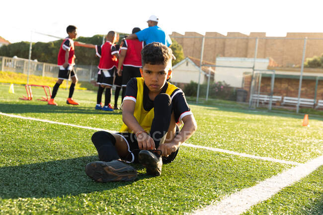 Vue de face d'un footballeur de race mixte assis sur un terrain de jeu au soleil mettant ses bottes de football lors d'une séance d'entraînement, ses coéquipiers écoutant leur entraîneur en arrière-plan — Photo de stock