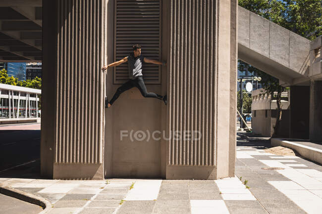 Vista lateral de um homem caucasiano praticando parkour perto do edifício em uma cidade em um dia ensolarado, escalando parede de concreto de um edifício moderno. — Fotografia de Stock