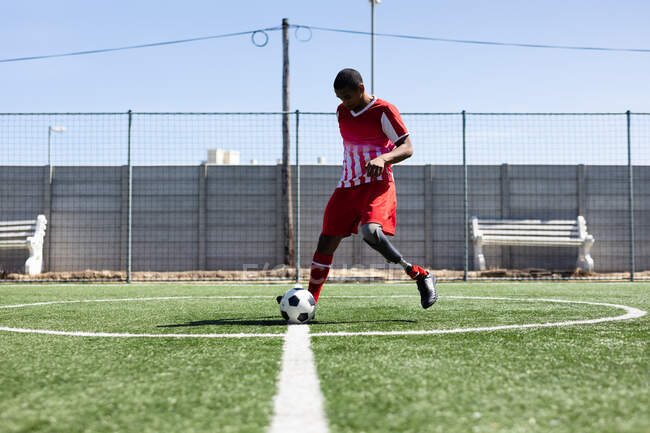 Giocatore di calcio maschile di razza mista con gamba protesica che indossa un allenamento di striscia di squadra in un campo sportivo al sole, scaldando la palla. — Foto stock