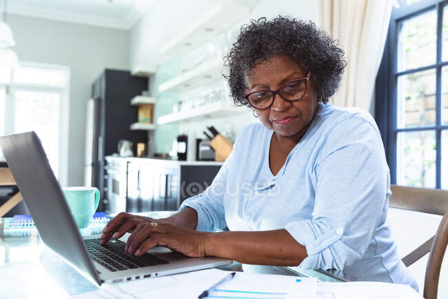 Mujer de raza mixta mayor disfrutando de su tiempo en casa, distanciamiento social y aislamiento en cuarentena, sentada en una mesa, usando una computadora portátil - foto de stock