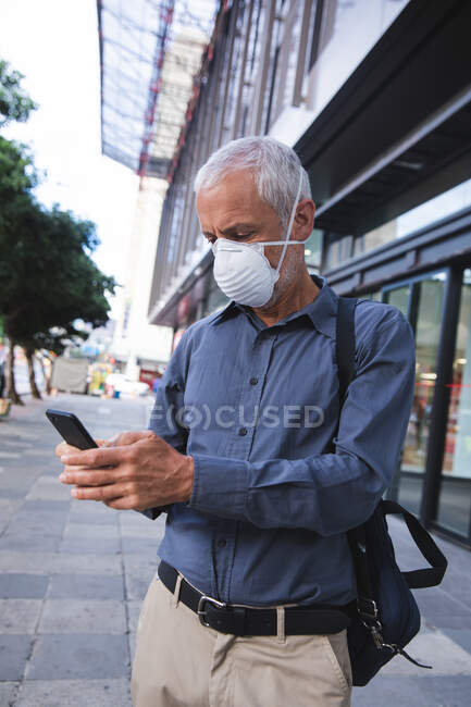 Hombre caucásico mayor por las calles de la ciudad durante el día, usando una máscara facial contra el coronavirus, covid 19 y usando un teléfono inteligente. - foto de stock