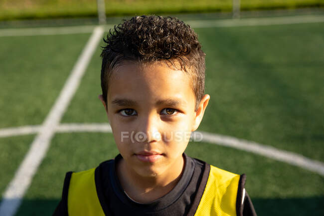 Портрет крупним планом впевнений змішаної раси хлопчик-футболіст в командній смузі, стоячи на ігровому полі на сонці, дивлячись прямо на камеру — стокове фото