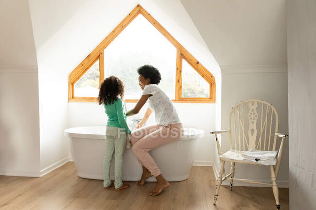 Vista lateral de una mujer afroamericana y su hija pequeña en el baño, la madre sentada en el borde de la bañera y corriendo un baño, la hija de pie junto a ella - foto de stock