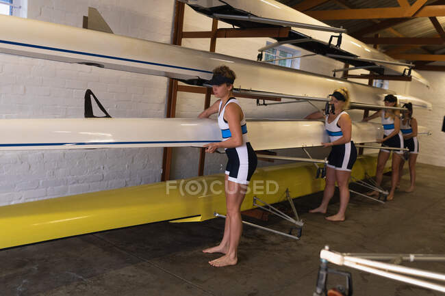 Side view of a rowing team of four Caucasian women lifting a boat from a storage rack in a boathouse — Stock Photo