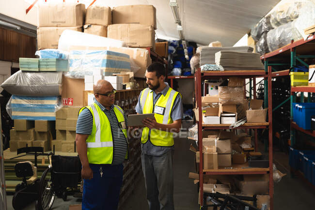 Mixed race and Caucasian male workers in a storage warehouse at a factory making wheelchairs, standing and using tablet, wearing a workwear — Stock Photo