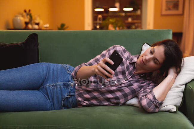 Смешанная расовая женщина проводит время дома самоизолируясь и социальное дистанцирование в карантинной изоляции во время эпидемии коронавируса ковид 19, лежа на диване, используя смартфон в гостиной. — стоковое фото