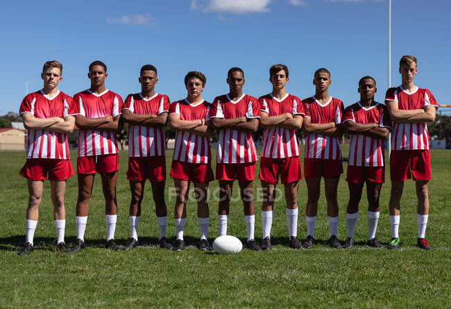 Frontansicht einer Gruppe multiethnischer männlicher Rugby-Spieler mit rot-weißer Mannschaftskleidung, die mit verschränkten Armen auf einem Spielfeld stehen und in die Kamera schauen, während der Ball vor ihnen auf dem Boden liegt — Stockfoto