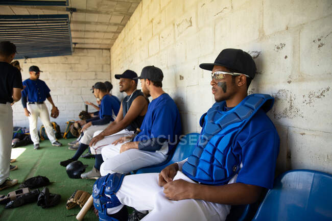 Vista lateral de una fila de jugadores de béisbol masculinos multiétnicos, preparándose antes de un juego, sentados en el vestuario, enfocándose mientras esperan, interactuando - foto de stock