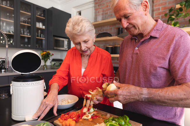 Heureux couple caucasien âgé à la retraite à la maison, préparant la nourriture et souriant dans leur cuisine, l'homme coupant des légumes, la femme regardant et lui parlant, à la maison ensemble isolant pendant la pandémie de coronavirus covid19 — Photo de stock
