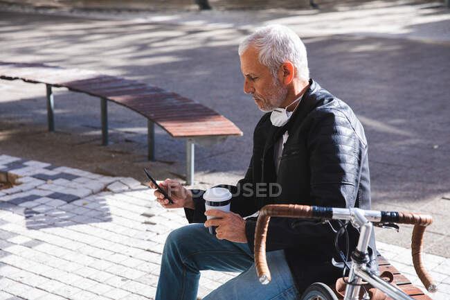 Uomo anziano caucasico in giro per le strade della città durante il giorno, indossando una maschera contro il coronavirus, covid 19, seduto su una panchina, tenendo una tazza di caffè da asporto e usando il suo smartphone. — Foto stock
