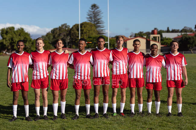 Vista frontal de un grupo de jugadores de rugby masculinos multiétnicos adolescentes que usan tira de equipo roja y blanca, de pie en un campo de juego, abrazándose. - foto de stock