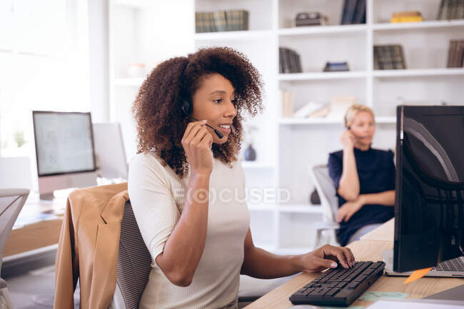 Eine Geschäftsfrau mit gemischter Rasse, die in einem modernen Büro arbeitet, am Schreibtisch sitzt, einen Computer benutzt, ein Headset trägt und telefoniert, während ihr Geschäftspartner im Hintergrund arbeitet. — Stockfoto