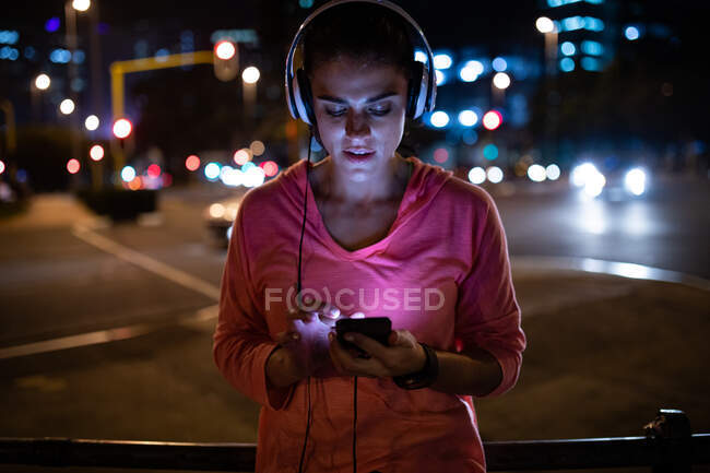 Front view of a fit Caucasian woman with long dark hair wearing sportswear exercising outdoors in the city in the evening, taking a break from her workout standing with headphones using a smartphone with urban buildings in the background. — Stock Photo