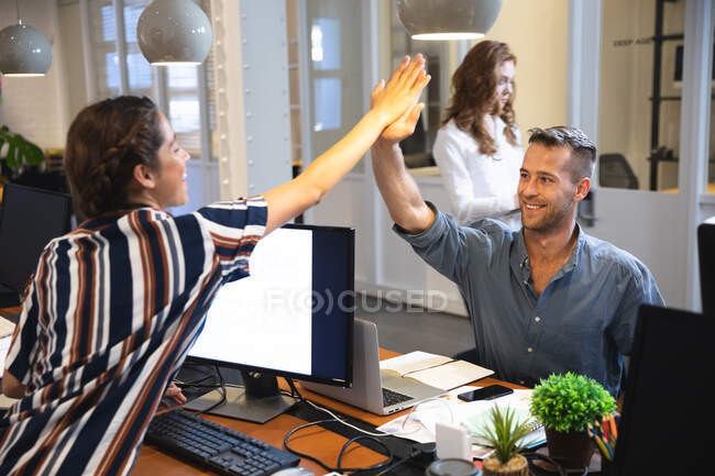 Creativos de negocios masculinos femeninos y caucásicos de raza mixta felices que trabajan en una oficina moderna informal, sentados en una mesa, chocando contra las pantallas de su computadora y sonriendo - foto de stock