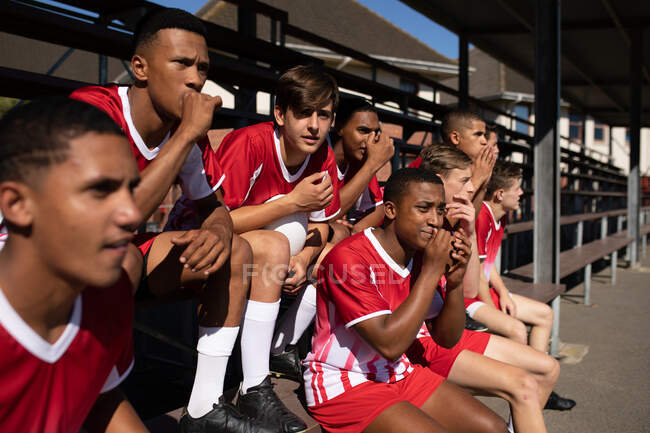 Vue latérale d'un groupe de joueurs de rugby masculins multi-ethniques adolescents portant une bande rouge et blanche, acclamant depuis les gradins lors d'un match — Photo de stock