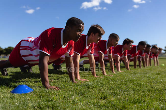 Vista laterale di una squadra maschile multietnica adolescente di giocatori di rugby che indossano la loro striscia di squadra, facendo press up sul campo di gioco di fila — Foto stock