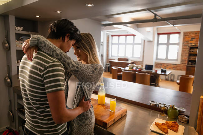 Vista lateral de um jovem mestiço e uma jovem caucasiana de pé na cozinha, abraçando-se enquanto preparavam o café da manhã juntos . — Fotografia de Stock