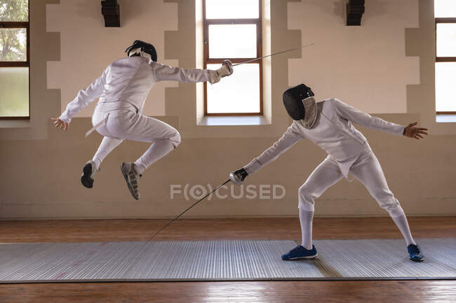 Caucasian and mixed race sportsmen wearing protective fencing outfits during a fencing training session jumping while duelling with their epees. Fencers training at a gym. — Stock Photo