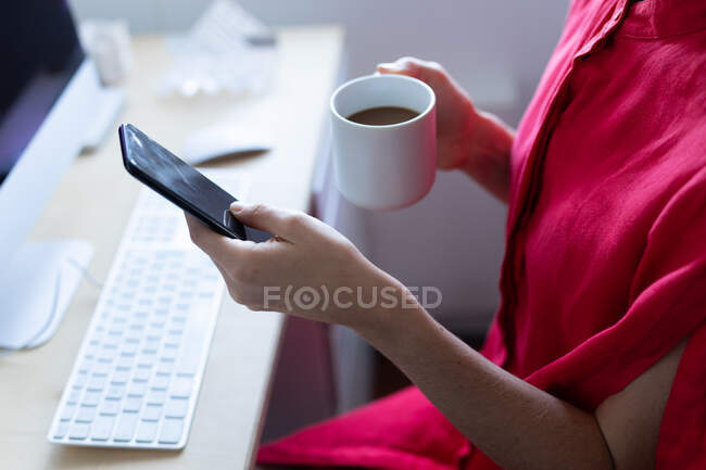 Vista a meio da seção da mulher passar o tempo em casa, vestindo um vestido rosa, sentado ao lado de sua mesa, segurando uma xícara de café e usando seu smartphone. Distanciamento social e auto-isolamento em quarentena . — Fotografia de Stock