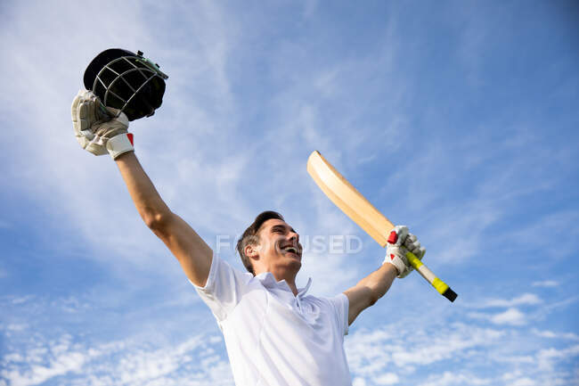 Visão lateral de baixo ângulo de um jogador de críquete masculino caucasiano adolescente vestindo brancos, em pé no campo, sorrindo e levantando as mãos, segurando um taco de críquete e um capacete de críquete. — Fotografia de Stock