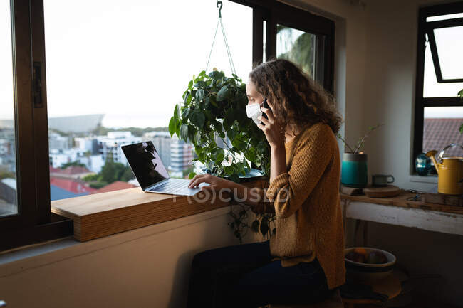 Белая женщина проводит время дома, изолируя себя, надевая маску для лица против вируса ковид19, стоя у окна, разговаривая на смартфоне и работая с ноутбуком. — стоковое фото