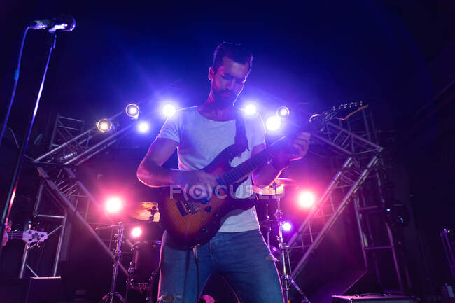 Vista frontal de cerca de un guitarrista caucásico centrado en tocar su guitarra eléctrica, de pie en un escenario iluminado con una banda actuando en un lugar de música, con un bajista y un kit de batería solo visible en el fondo - foto de stock