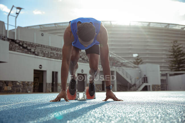 Front view of a mixed race male athlete practicing at a sports stadium, in position on starting blocks, preparing to sprint, with his head down, backlit by sunlight — Stock Photo