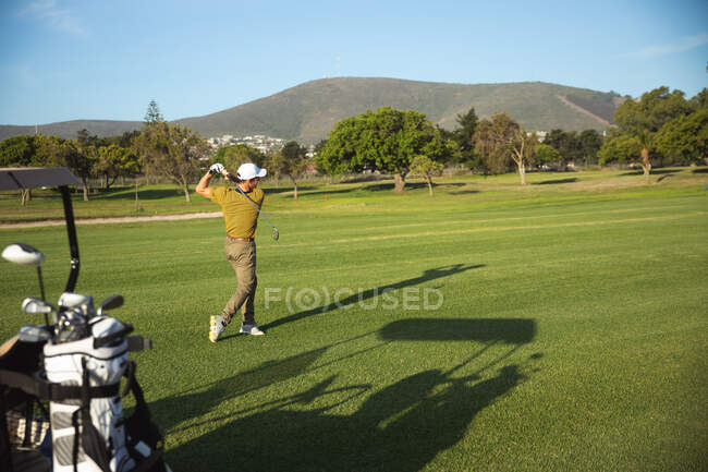 Vista lateral de un hombre caucásico en un campo de golf en un día soleado con cielo azul, golpeando una pelota de golf - foto de stock
