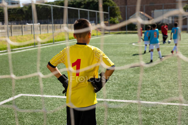 Задний план кавказский мальчик, стоящий в воротах с руками на бедрах, во время игры между двумя многонациональными командами мальчиков футболистов носить их командные полоски, в действии во время футбольного матча на игровом поле — стоковое фото