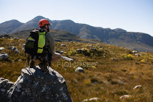 Vista trasera del hombre caucásico disfrutando del tiempo en la naturaleza, con equipo de forro de cremallera, senderismo, sentadillas en una roca en un día soleado en las montañas - foto de stock