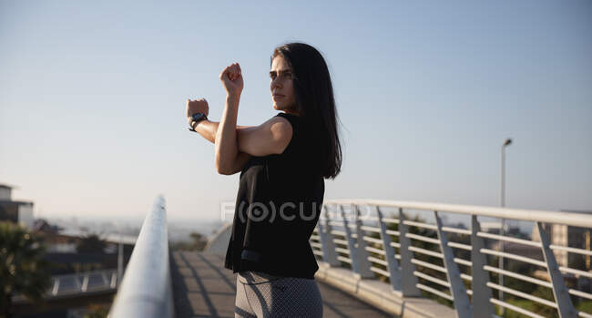 Seitenansicht einer fitten kaukasischen Frau mit langen dunklen Haaren, die an einem sonnigen Tag mit blauem Himmel draußen in der Stadt Sport treibt, sich aufwärmt und die Arme ausstreckt. — Stockfoto